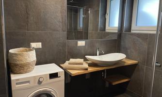 A modern bathroom with dark tiles and a sink on a wooden shelf. A washing machine and a laundry basket are also visible.