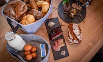 A basket with different types of bread is on a table, next to it are eggs, milk, and ham on a black board. The scene is cozy and inviting.