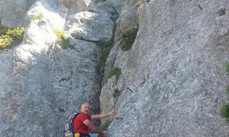 A climber is at a steep rock wall and using a safety gear. The sky is clear and the surroundings are green.