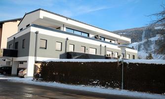 A modern residential building with several floors, surrounded by snow. Snow-covered mountains are visible in the background.