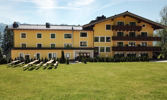 A cozy yellow hotel with a large garden and comfortable outdoor benches. In the background, green trees and mountains can be seen.