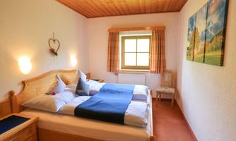 A cozy bedroom with two beds and a window. The walls are plain, and there is light wood along with decorations.