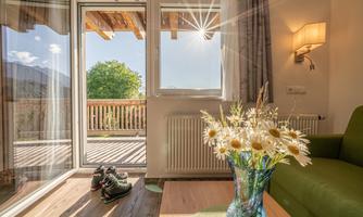 A cozy living room with a green sofa and a vase full of flowers. The sun is shining through the windows and you can see a terrace with a view of the outdoors.