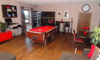 A cozy room with a billiard table in the center and modern furniture. There is a bar, small seating areas, and decorative plants.