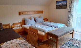 A cozy bedroom with two beds, a chair, and a table. The room has large windows that let in plenty of daylight.