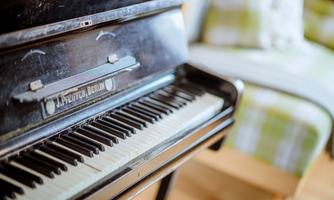 A black piano with visible keys and a glossy surface. In the background, there is a piece of seating furniture with a checked pattern.
