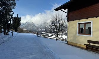A snowy path leads to a house in the mountains. The landscape is calm and picturesque, surrounded by snow-covered mountains and trees.