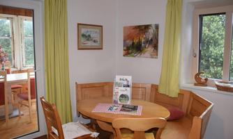 A cozy dining area with a round table and a bench. The walls are painted in light colors, and there are windows that let in plenty of light into the room.