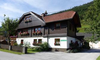 A charming house in alpine style with flower windows and a well-maintained garden. In the background, green mountains can be seen.