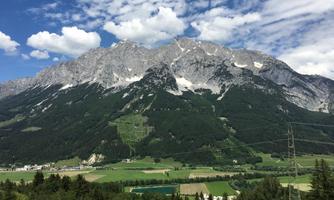 An impressive mountain landscape with high, snow-covered peaks and lush green forests in the foreground. The sky is blue with some white clouds.