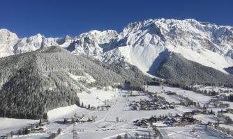 A snow-covered landscape with impressive mountains and a clear blue sky. In the foreground, there is a small village, surrounded by winter beauty.
