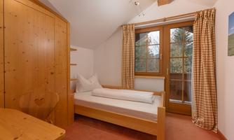 A cozy bedroom with a single bed and a wooden wardrobe. Large windows with curtains let in daylight.
