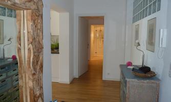 A bright hallway with a wooden mirror and a chest of drawers. In the background, another room with warm light is visible.