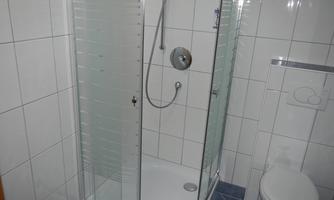 A modern shower with a glass shower partition and tiled floor. In the background, a toilet can be seen.