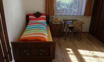 A cozy bedroom with a single bed and colorful cushions. A small table and chair complement the room, which receives plenty of daylight.