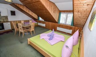 A cozy attic room with a double bed and a dining table. The room has a wooden ceiling and large windows that let in plenty of light.