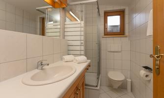 A modern bathroom with a sink, a shower cubicle, and a toilet seat. The walls are tiled, and there are windows for natural light.