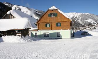 A charming wooden house in the snow with mountains in the background. The sky is blue and clear.