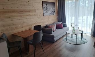 A cozy living area with a gray sofa and a wooden dining table. Large windows let in a lot of light, and the decoration is modern and inviting.