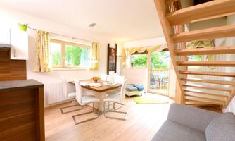 A bright and modern apartment with a dining area and large windows. A wooden staircase leads to additional rooms.