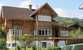 A beautifully designed wooden house with a balcony and large windows. Behind it, green hills and a clear blue sky can be seen.