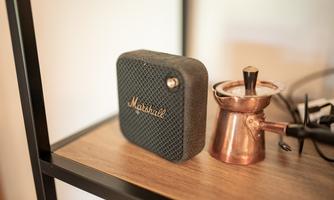 A black speaker from Marshall is sitting on a wooden table next to a copper coffee machine. The background is blurred and bright.