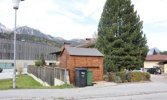 A wooden house stands next to a large tree. In the background, mountains can be seen, and there are trash cans in front of the house.