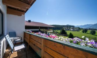 A beautiful balcony with blooming flowers and a view of the green meadows and mountains. The clear sky completes the idyllic scenery.