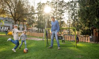 A father plays with his children in the garden. The children are playing soccer with a football, while the sun shines on the horizon.