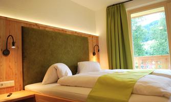 A cozy bedroom with a modern wooden bed and a green bedspread. Large windows provide plenty of daylight and a view outside.
