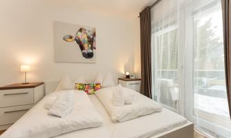 A cozy bedroom with a large bed and colorful cushions. A modern cow picture hangs on the wall, and large windows let in plenty of light.