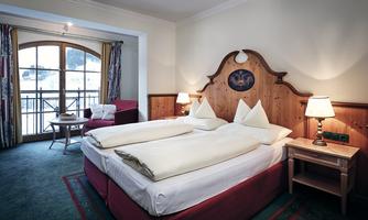 A cozy hotel room with two single beds, a red sofa, and a window with a view. The walls are painted in warm colors and there are nightstands with lamps.