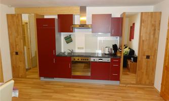 A modern kitchen with red cabinets and wooden accents. The floor is made of wood and the wall is painted light.