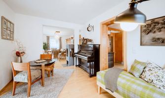 A cozy living room with a piano, a dining table, and a sofa. The bright walls and wooden floor create a pleasant atmosphere.