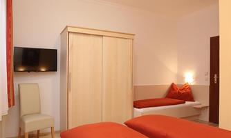 A simple and cozy room with two single beds and warm red bed covers. On the wall, there is a television and a wooden wardrobe.