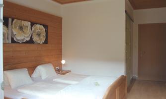 A simple room with two beds and a wooden wall design. Pictures of flowers hang on the wall, and there is a lamp on the nightstand.