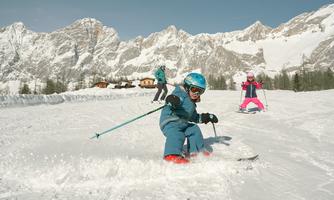 A boy is skiing fast in the snow, while two other children are skiing in the background. The surroundings are surrounded by snow-covered mountains and the sky is clear.