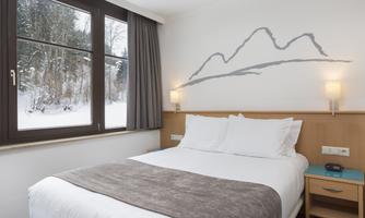A cozy hotel room with a comfortable bed and a bright window front. Outside, a snowy landscape is visible.