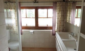 A bright bathroom with a window, shower, and modern sanitary facilities. The walls are tiled and decorated with curtains.
