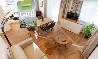 A modern living room with a dining area, a TV corner, and an elegant table. The room is bright and friendly, with wooden flooring and large windows.