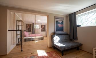 A cozy room with a sleeping area in the background and a comfortable armchair in the foreground. Large windows provide plenty of daylight.
