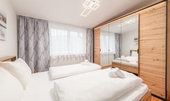 A modern bedroom with a double bed and two white duvets. Against the wall stands a large wardrobe with a mirror and light curtains.