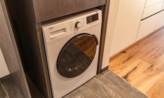 A modern washing machine in a kitchen unit. The floor is designed with dark tiles and wooden coverings.