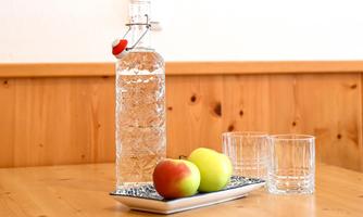 A glass bottle of water is on a table next to two apples. Next to it are two glasses on a plate.