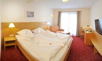 A cozy hotel room with a large bed, a seating area, and plenty of natural light. The walls are bright, and the decor is simple and modern.