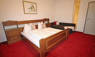 A cozy bedroom with a wooden bed, two pillows, and red carpet flooring. A picture hangs on the wall, and there is an additional sofa bed.