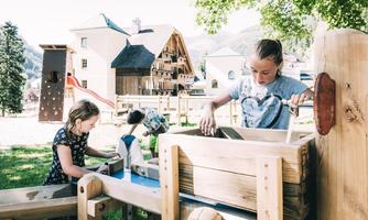 Children are playing at a water playground with wooden structures. In the background, houses and trees can be seen.