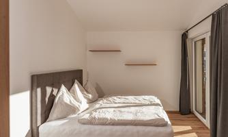 A bright bedroom with a spacious bed and wooden floor. Large windows provide plenty of daylight and a pleasant atmosphere.