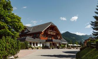 A picturesque hotel in alpine style, surrounded by trees and mountains. The sky is clear and blue, perfect for a relaxing getaway.