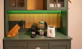 A cozy kitchenette with a coffee machine, a kettle, and a toaster. The green lighting creates a pleasant atmosphere.